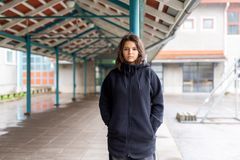 Tyttö nojaa ulkona pylvääseen ja katsoo suoraan kameraan vakavana. Taustalla asfalttia ja katos