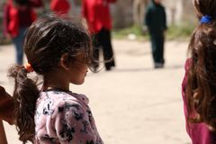 Tyttö seisoo ulkona, hänellä on poninhäntä ja päällään kuviollinen paita. Taustalla useita ihmisiä.