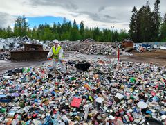 Ympäristöasiantuntija lapioi metallijätettä kierrätyskeskuksessa, jossa näkyy paljon erilaisia metallipakkauksia ja pienmetallia.