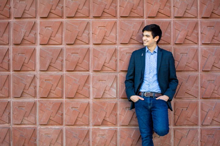 Ystävällinen mies nojaa tiiliseinään, hymyilee ja katsoo sivulle. Hänellä on päällään puku ja farkut.