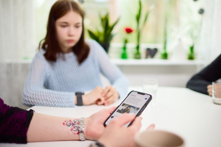 Nuori lapsi katsoo pöydän ääressä, kun viereinen henkilö käyttää puhelintaan.