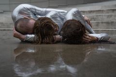 Kolme vaaleisiin, kiiltäviin ja ihonmyötäisiin vaatteisiin pukeutunutta pääpuolelta kuvattua ihmistä makaa sylikkäin lattialla kivirappusten edessä. Kenenkään kasvoja ei näy.