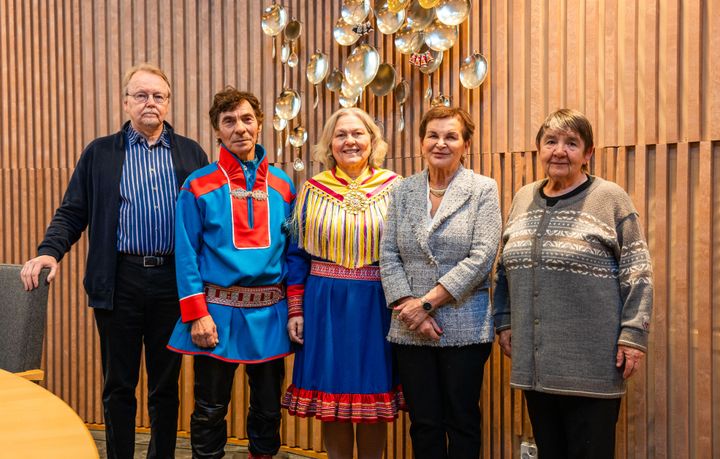 Viisi henkilöä ryhmäkuvassa, vasemmalla tummapukuinen mies, vieressä saamenpukuinen mies, hänen vieressään keskimmäisenä kuvassa saamenpukuinen vaaleahiuksinen nainen. Oikealla laidalla kaksi lyhyt-tummahiuksista naista. Takana kaunis puuseinä ja seinällä saamelaista korua, riskua muistuttava iso taideteos. Henkilöt hymyilevät.